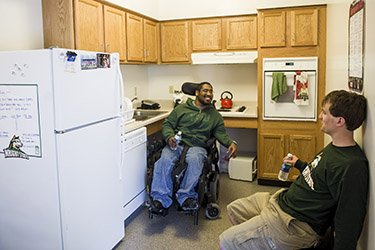Two student are in an accessible apartment kitchen, one is seated in a power wheelchair the other on a chair