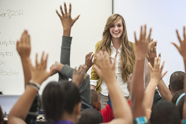 Teacher in front of excited children raising their hands to her 