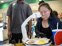 A white robotic arm brings a spoon to the mouth of a young woman.  There are a variety of foods in the 4 sections of the plate for the diner to choose from. 